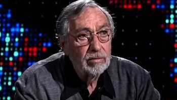 Primer plano de Luis Brandoni, un hombre mayor con pelo y barba grises, gafas redondas, y una camisa oscura, mirando pensativo