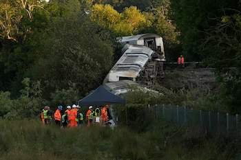 Vagones de un tren descarrilado entre la vegetación, con personal de emergencia, una tienda de campaña oscura y la vía férrea