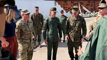 Varios militares en uniforme y una mujer civil conversan al aire libre con una pared parcialmente derrumbada y escombros de fondo