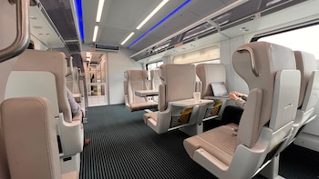 Interior de un tren Brightline: asientos beige, pasajero con laptop en mesa desplegable y vaso de café, ambiente moderno