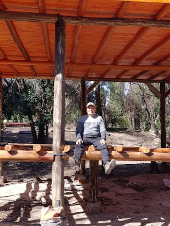 Hombre con gorra y jersey bicolor sentado sobre una viga de madera rústica. La estructura tiene un techo de madera y, al fondo, se aprecian árboles