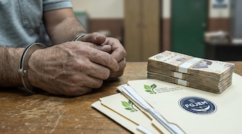 Primer plano de manos masculinas esposadas en una mesa. Hay billetes mexicanos de 200 pesos y documentos de la Secretaría del Medio Ambiente y FGJEM.