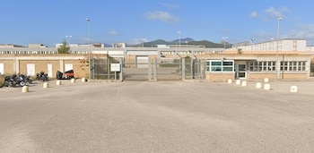 Entrada exterior de un complejo penitenciario con caseta de vigilancia, portón de rejas, aparcamiento con motocicletas y colinas de fondo