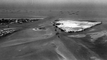 Fotografía aérea en blanco y negro de una zona costera con una playa prominente, agua y múltiples barcos. En el mar distante hay varios buques