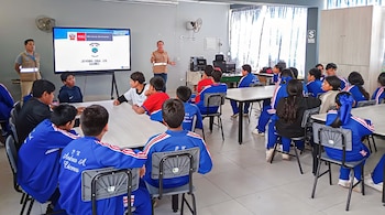 Varios escolares con uniforme azul se sientan ante mesas en un aula mientras personal de la Sucamec expone frente a una pantalla