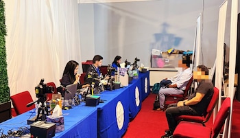 Vista de un equipo de producción de medios trabajando en una mesa azul con cámaras y laptops. Varias personas sentadas en sillas rojas, algunas de espaldas