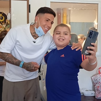 Enzo Fernández, con camiseta blanca y mascarilla en la barbilla, posa junto a un niño de camiseta azul que sostiene un teléfono móvil