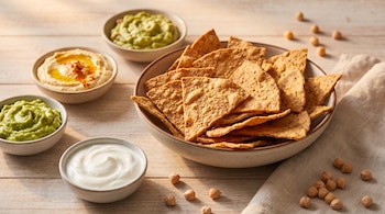Un tazón de nachos de garbanzo. Hummus, dos tazones de guacamole y uno de yogur griego en recipientes. Garbanzos sueltos y servilleta en mesa de madera.
