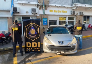 Dos agentes de la DDI con uniforme negro, un coche Peugeot 207 plateado, un cartel de la DDI y el edificio de la DDI Mar del Plata