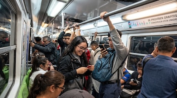 Personas de pie y sentadas dentro de un vagón de tren subterráneo con pasamanos superiores, ventanas, asientos verdes y carteles informativos.