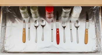 Cajón de madera con base de papel aluminio brillante. Contiene frascos de especias y utensilios de cocina metálicos y con mangos de madera y rojos, organizados.