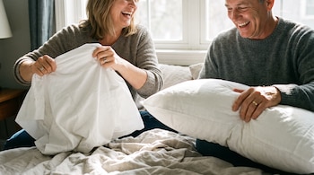 Hombre y mujer sentados en la cama; la mujer sujeta una funda de almohada blanca, el hombre sostiene una almohada blanca. Ventana detrás.