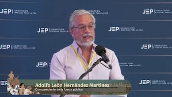 Hombre con barba, gafas, camisa rosa, y acreditación amarilla, hablando en micrófono. Fondo con logos de JEP Jurisdicción Especial para la Paz. Gráfico con su nombre y cargo