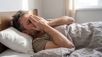 Hombre recostado en una cama con almohada blanca y manta gris que cubre su rostro con las manos.