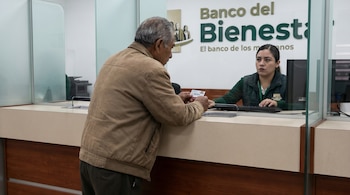 Hombre mayor con chaqueta beige se inclina en un mostrador de banco, mostrando una tarjeta. Una cajera de uniforme verde lo atiende detrás de una mampara de cristal. El logo del Banco del Bienestar es visible al fondo.