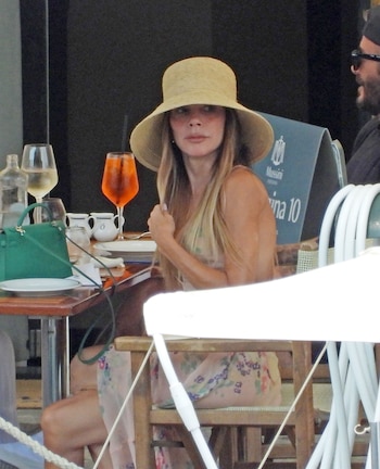 Mujer con sombrero de paja y vestido floral sentada en una mesa. Hay una copa de vino blanco, un cóctel naranja, una botella de agua y un bolso verde