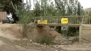 Puente de metal sobre un cauce de tierra rodeado de cañas y vegetación, junto a un camino no asfaltado y una motocicleta estacionada
