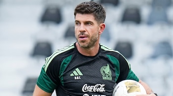 Hombre con camiseta negra y verde del equipo de México, con logotipos de patrocinadores. Sostiene un balón de fútbol blanco y dorado. Fondo borroso.