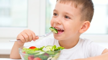 Un niño sostiene un tenedor con una rodaja de pepino frente a un cuenco transparente con ensalada, lechuga y tomates.