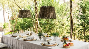 Mesa de comedor exterior con mantel blanco, servicios de mesa, cuencos de sopa, ensalada y verduras. Dos lámparas de mimbre cuelgan con árboles de fondo.