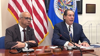 Dos hombres en trajes sentados a una mesa de madera con micrófonos y banderas de Estados Unidos y la OEA de fondo. Uno habla, el otro escucha atentamente