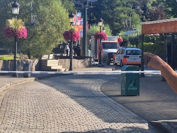 Calle adoquinada con cinta de seguridad. Farolas con flores, cubo de basura Plopsa, árboles, furgoneta, coche y una mano señalando