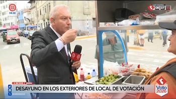Un hombre de traje come de un cuenco pequeño mientras sostiene un micrófono frente a un puesto de comida callejera con ceviche y limones