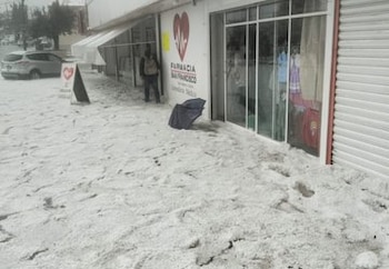 Vista exterior de una farmacia con sus alrededores cubiertos por una gruesa capa de granizo. Un hombre, un auto y una sombrilla rota son visibles