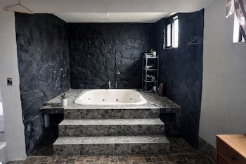 Bañera blanca de hidromasaje elevada con tres escalones de azulejos, paredes oscuras de piedra, suelo de mosaico, ventana lateral, estantería metálica y planta.