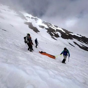 Tres personas transportan una camilla naranja sobre la nieve en una ladera. Al fondo, una montaña con nieve y rocas bajo un cielo nublado