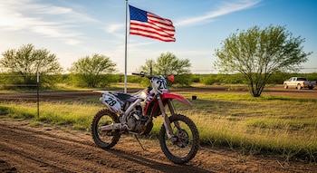 Una moto de motocross roja y blanca estacionada en un camino de tierra, con una bandera de Estados Unidos ondeando en un poste y árboles en un paisaje rural.