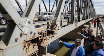 Grupo de vecinos señala la sección de un puente con pernos faltantes y corrosión, mientras vehículos transitan por la calzada y un río fluye debajo.
