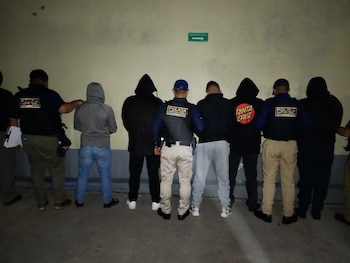 Un agente con chaleco de inspector camina junto a una persona con capucha negra en una acera durante la noche