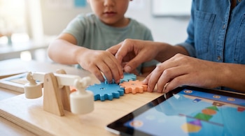 Manos de un niño y una docente manipulando engranajes de juguete, una balanza didáctica y una tableta digital sobre una mesa de madera.