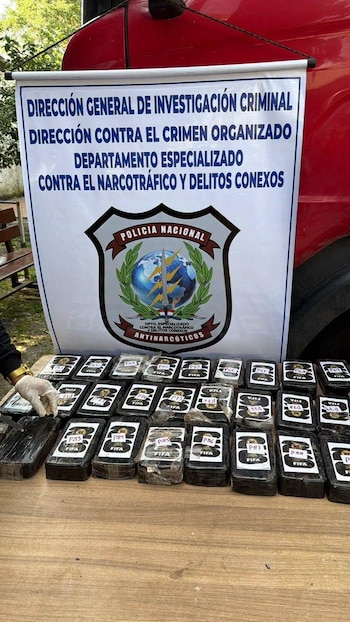 Mesa de madera con paquetes rectangulares negros, algunos con inscripciones 'FIFA' y códigos alfanuméricos, junto a un cartel de la Policía Nacional