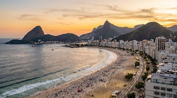 Vista panorámica de una playa con personas, edificios a lo largo de la costa, el mar y montañas en el horizonte bajo un cielo al atardecer.