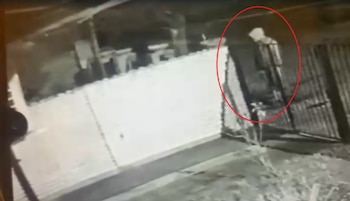 Imagen granulada de cámara de seguridad nocturna en blanco y negro mostrando a una persona con capucha cerca de una valla. Un círculo rojo la resalta