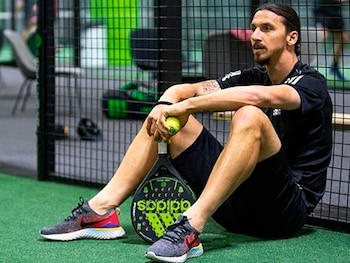 Un hombre de cabello largo y barba sentado en el suelo sintético de una pista de pádel con una pala y una pelota