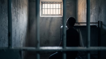 Silueta de un hombre de espaldas sentado en una celda de hormigón con una litera metálica y una ventana con barrotes.