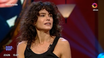 Mujer de cabello rizado oscuro y camiseta negra en un estudio de televisión con logotipos y texto en pantalla