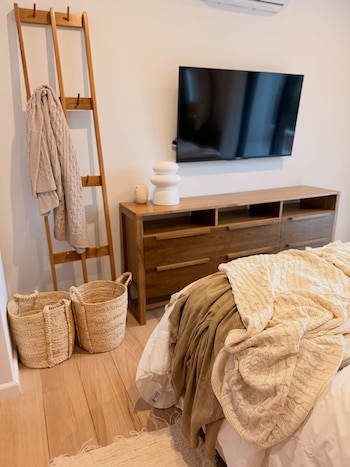 Dormitorio moderno con cómoda de madera, TV, perchero escalera con manta beige, dos cestas de mimbre en el suelo de parquet y cama con textiles neutros