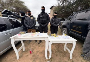 Cuatro agentes de seguridad uniformados parados detrás de dos mesas blancas con objetos incautados, junto a dos vehículos al aire libre