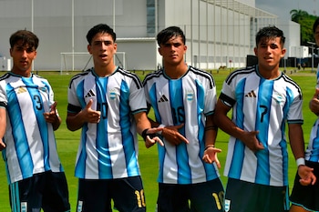 Cuatro jóvenes futbolistas argentinos con camisetas a rayas celestes y blancas posan en un campo de fútbol con un edificio blanco de fondo