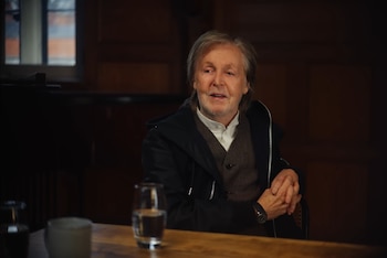 Paul McCartney, con cabello gris y barba, sentado en una mesa de madera. Viste chaqueta oscura, camisa blanca y chaleco. Manos entrelazadas. Vaso de agua en primer plano