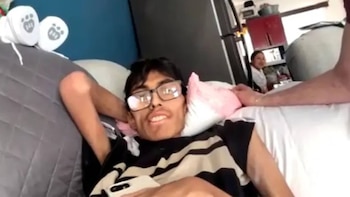 Un joven con gafas, cabello oscuro y camiseta a rayas se recuesta en un sofá gris, sosteniendo un teléfono blanco. Una almohada rosa a su lado