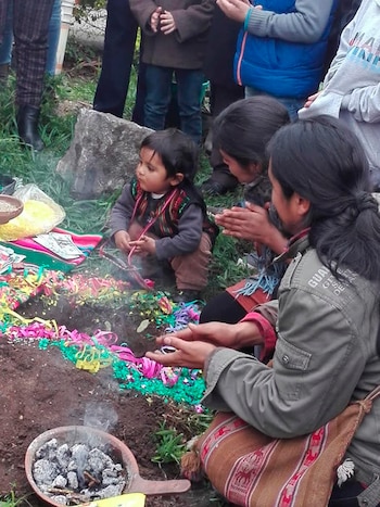 Varias personas, incluyendo un niño, se congregan alrededor de un pozo en la tierra decorado con cintas de colores. Un recipiente con brasas humeantes está en primer plano