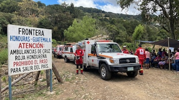 Un cartel grande marca la frontera entre Honduras y Guatemala, indicando que el paso está prohibido para ambulancias de la Cruz Roja Hondureña. Hay ambulancias y personal con uniformes rojos, con personas esperando bajo una lona al fondo.