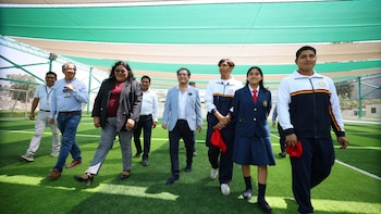 Un grupo de adultos y estudiantes camina sobre un campo de césped artificial bajo un techo de tela verde y blanca