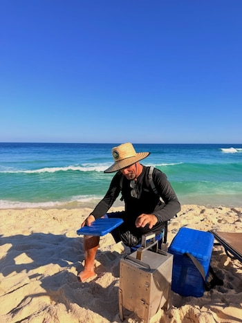 Um homem de chapéu de palha e camiseta preta está sentado na areia. Mar azul, céu limpo, equipamentos metálicos e geladeira azul na praia