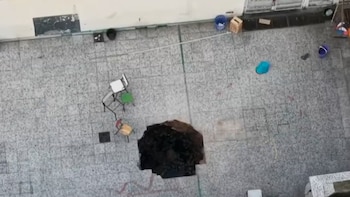 Vista desde arriba de un patio de baldosas grises con un gran orificio en el piso, rodeado por sillas y baldes de plástico